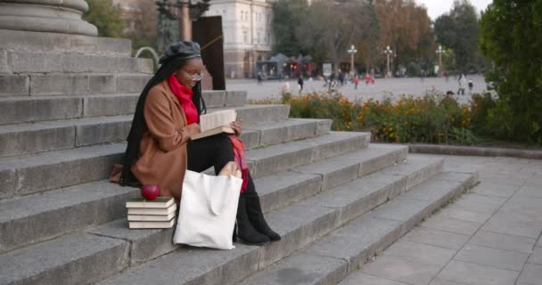 Une jeune fille noire est assise dans les escaliers dans la rue et lit un livre. — Video