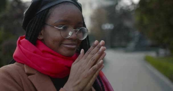 Jong zwart meisje in een jas en sjaal verwarmt haar handen — Stockvideo