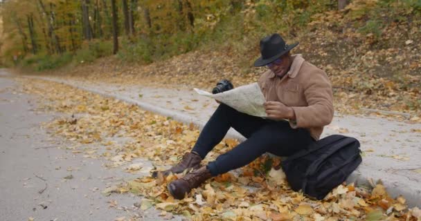 Ένας μαύρος ταξιδιώτης κάθεται στην άκρη του δρόμου με ένα χάρτη — Αρχείο Βίντεο