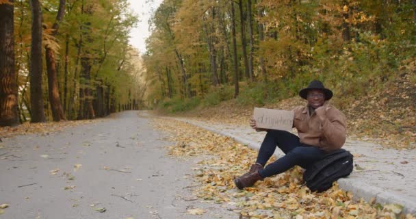 Молодой черный автостопщик с табличкой на обочине дороги — стоковое видео
