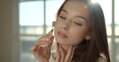 Evde yüz masajı için yeşim yüz silindiri kullanan kadın.