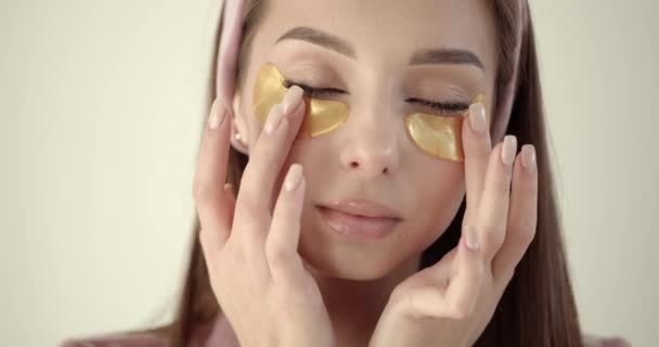 Woman applying hydrogel under-eye recovery patches on a light background — Video
