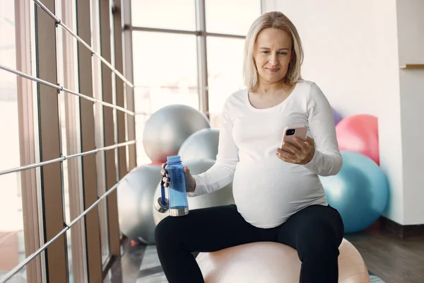 Pregnant woman using a phone while sitting in bright fitness studio — Fotografie, imagine de stoc