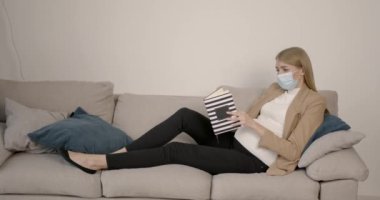 Maskeli hamile kadın kanepede kitap okuyor.