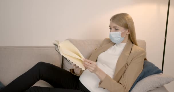 Mulher grávida em máscara livro de leitura no sofá — Vídeo de Stock