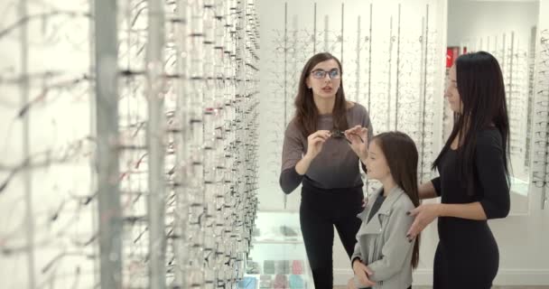 父母们带着可爱的女儿在一家现代光学商店里试戴眼镜 — 图库视频影像