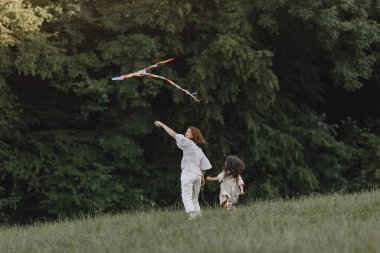 Anne ve kızı güneşli çayırda uçurtmayla oynuyorlar.
