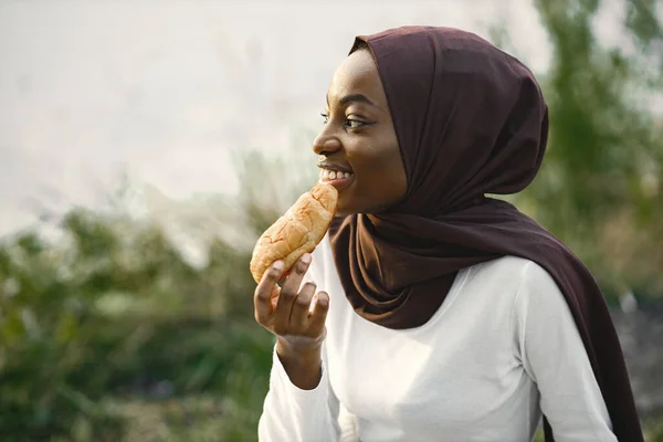 Portrait of a muslim woman sitting near the river and eating croissant — Stock Photo, Image
