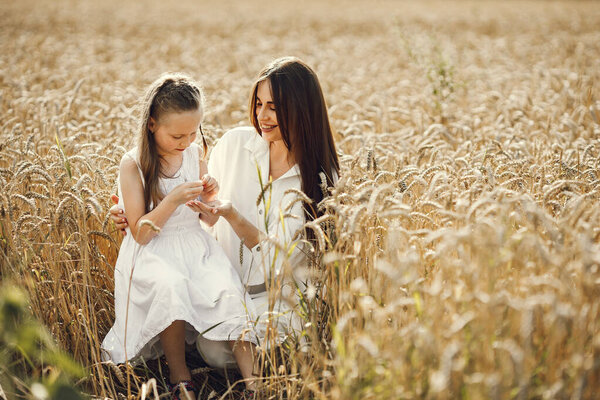 ute photo of a young mother and her daughter in white dresses at the wheat field on a sunny day. Brunette mother and her little daughter posing for a photo. Girl looking at wheat spike.