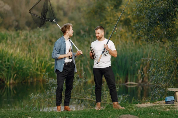 Friends is standing near the river and fishing. Two fishermen talking and drinking a beer. One man wearing white t-shirt and other blue shirt.