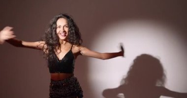 Çekici, kıvırcık saçlı, stil sahibi bir kadının eğlenmesi, dans etmesi, Noel partisinin tadını çıkarması, ellerini kaldırıp gülümsemesi, kendi vücudunun tadını çıkarması, ritme ayak uydurması, karanlık arka plana geçmesi..