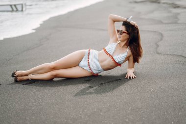 Esmer kız, Bali Endonezya 'da siyah kumsalda uzanmış güzel bir mayo giyiyor.