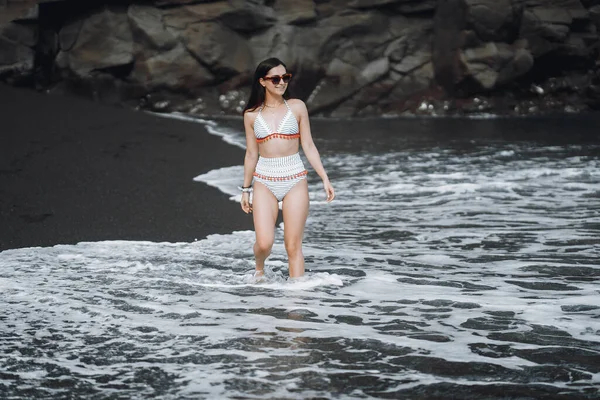黒砂のビーチを歩くブルネットの女の子は バリでかわいい水着を着てインドネシア — ストック写真
