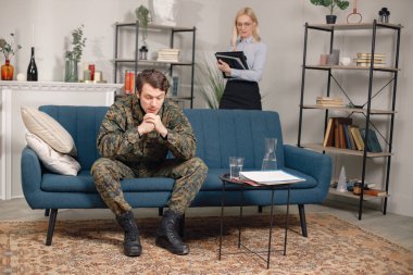 Askeri üniformalı bir adam kanepede oturuyor, kadın psikiyatrist arkada duruyor. Asker ve psikiyatrist terapi sırasında konuşuyor. PTD 'si olan erkek savaşçı zihinsel bozukluğunu tedavi ediyor..