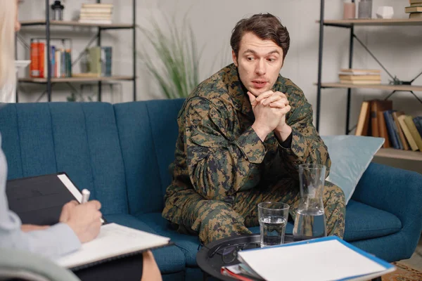 Cropped photo of a soldier and psychiatrist sitting on couch during therapy session. Man wearing military uniform. Male warrior with ptds talking to psychiatrist.
