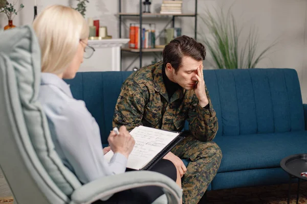 Soldier and psychiatrist sitting on couch during therapy session. Man wearing military uniform. Male warrior with ptds talking to psychiatrist.