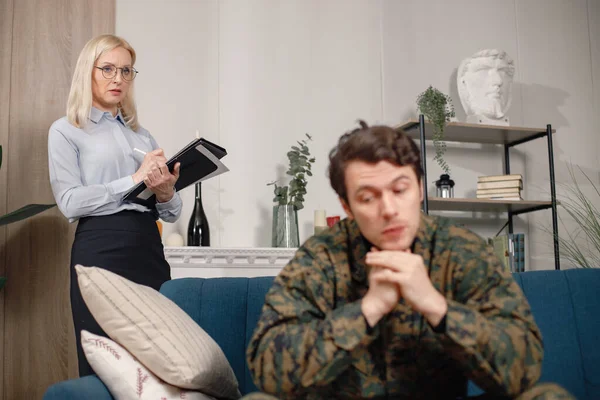 Man in military uniform sitting on a couch, woman psychiatrist standing behind. Soldier and psychiatrist talking during therapy session. Male warrior with ptds treating his mental disorder.
