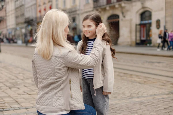 Genç anne kızını kucaklıyor. Kız yanağına Ukrayna bayrağını mavi ve sarı renklerle boyamış. Kadın ve kız Ukrayna 'yı savaşa karşı destekliyor.