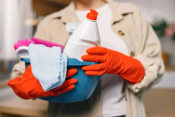 Cropped photo of a woman cleaning in a kitchen and wearing red gloves. Adult woman holding blue busket with different rugs and detergents. Cleaning concept.
