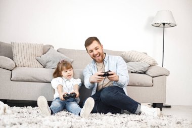 Sevgi dolu baba, Down sendromlu kızıyla evde birlikte oynuyor. Kız ve erkek kanepede oturup joystickleri tutuyorlar. Sakallı adam mavi gömlekli ve kız beyaz gömlekli..