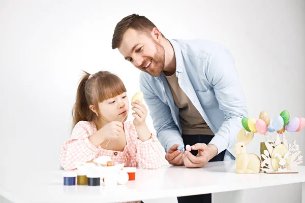 Portrait of a charming girl with Down syndrome wearing rose shirt and her bearded father. Girl sitting at desk in the kitchen playing with Easter colored eggs. Girl and her father preparing Easter