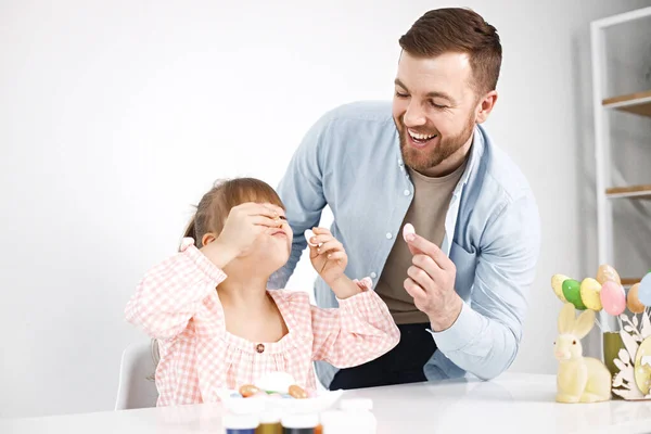 Portrait of a charming girl with Down syndrome wearing rose shirt and her bearded father. Girl sitting at desk in the kitchen playing with Easter colored eggs. Girl and her father preparing Easter