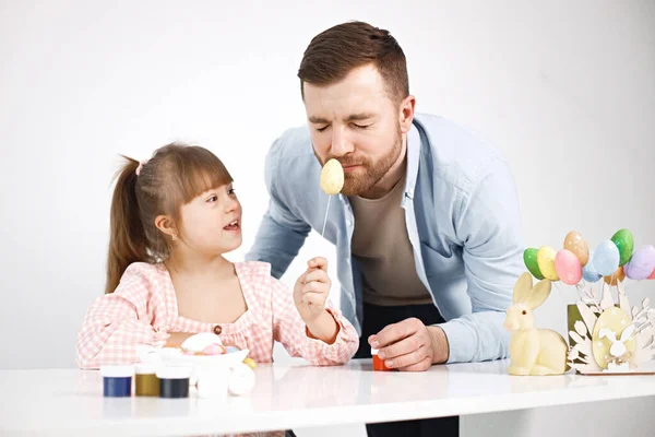 Portrait of a charming girl with Down syndrome wearing rose shirt and her bearded father. Girl sitting at desk in the kitchen playing with Easter colored eggs. Girl and her father preparing Easter