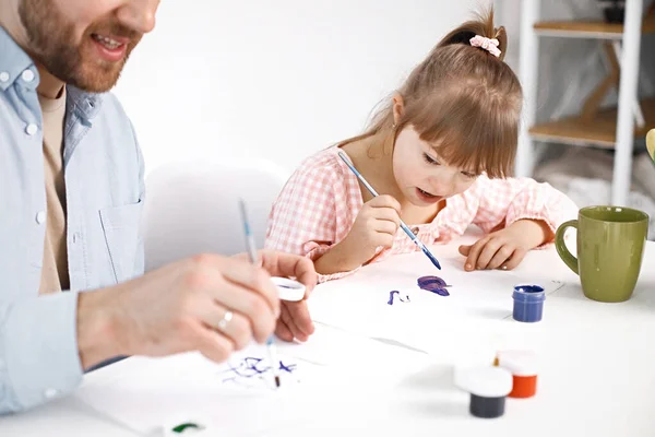 Portrait of a charming girl with Down syndrome wearing rose shirt and her bearded father. Girl sitting at desk in the kitchen painting Easter colored eggs. Girl and her father preparing Easter