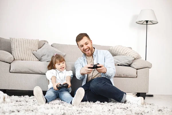 Loving father playing with his daughter with Down syndrome at home together. Man and girl sitting on a floor near sofa and holding joysticks. Bearded man wearing blue shirt and girl white shirt.
