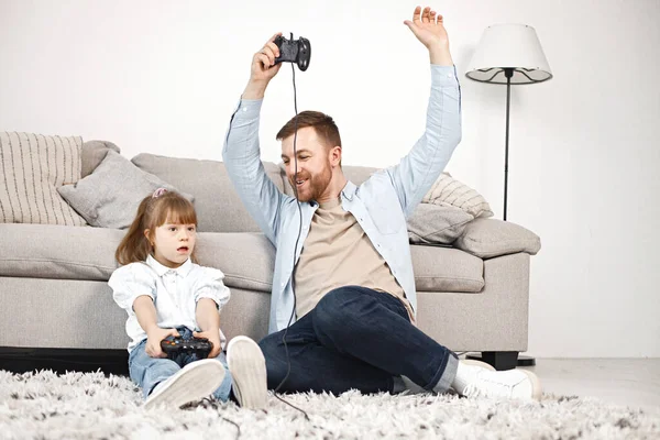 Sevgi dolu baba, Down sendromlu kızıyla evde birlikte oynuyor. Kız ve erkek kanepede oturup joystickleri tutuyorlar. Sakallı adam mavi gömlekli ve kız beyaz gömlekli..