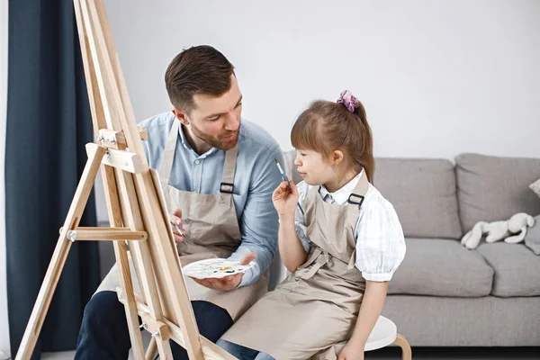 Little girl with Down syndrome wearing white shirt and beige apron. Girl with her father is painting on easel together. Father helping his daughter to paint.