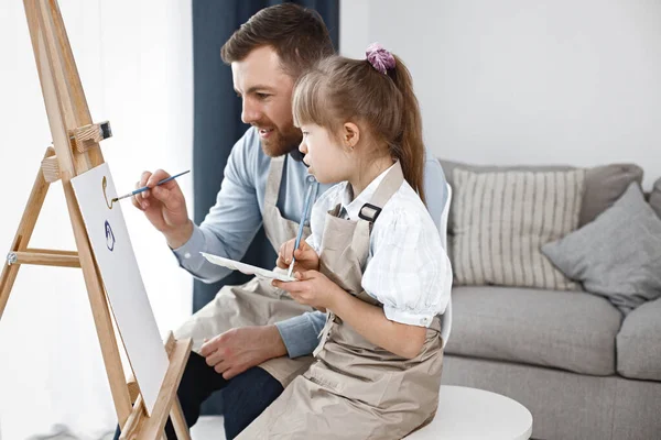 Little girl with Down syndrome wearing white shirt and beige apron. Girl with her father is painting on easel together. Father helping his daughter to paint.