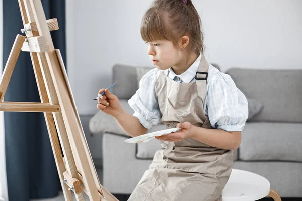 Little girl with Down syndrome wearing white shirt and beige apron. Girl is painting on easel. Girl sitting at home in living room.