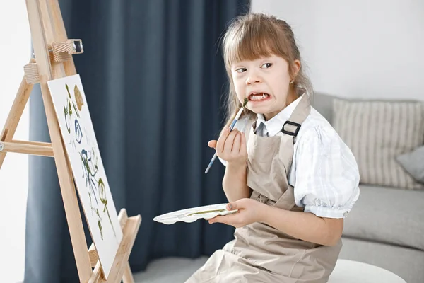 Little girl with Down syndrome wearing white shirt and beige apron. Girl is painting on easel. Girl sitting at home in living room.