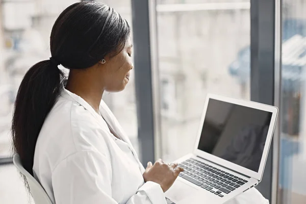 Young female black doctor sitting in her office at clinic near big windows. Woman wearing white medical coat. Brunette medical worker using a laptop.