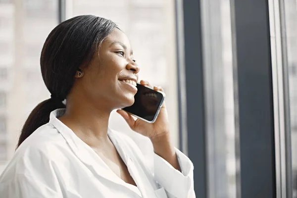 Young female black doctor standing in her office at clinic near big windows. Woman wearing white medical coat. Brunette medical worker using a phone.