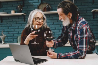 Yaşlı çift, ev mutfağında şarap içerken dizüstü bilgisayara bakmakla meşgul. Heyecanlı yaşlı erkek ve kadın sabah rutini sırasında bilgisayar kullanıyor. Yaşlı eş, bilgisayar başında kocasına bir şey gösteriyor..