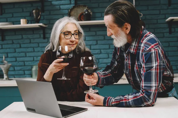 Aged couple busy look at laptop screen while drinking wine t at home kitchen, excited senior man and woman use computer during morning routine, elderly wife show something to husband at pc.