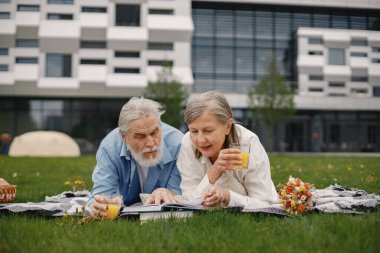 Yazın çimlerin üzerinde battaniyeye uzanmış beyaz yaşlı bir çift. Erkek ve kadın kitap okuyor ve meyve suyu içiyor. Beyaz tişörtlü kadın ve mavi erkek..