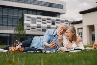 Yazın çimlerin üzerinde battaniyeye uzanmış beyaz yaşlı bir çift. Kadın ve erkek telefona bakıyor. Beyaz tişörtlü kadın ve mavi erkek..