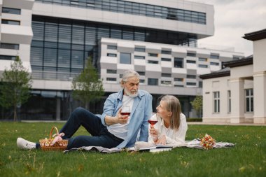 Yazın çimlerin üzerinde battaniyenin üzerinde oturan beyaz yaşlı bir çift. Kadın ve erkek şarap içiyor. Beyaz tişörtlü kadın ve mavi erkek..