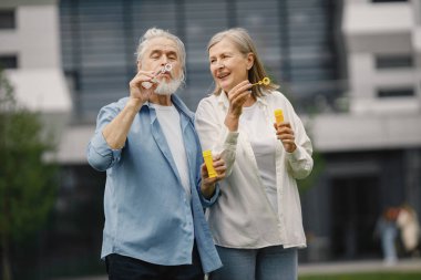 Yazın çimlerin üzerinde duran Kafkasyalı yaşlı çift. Erkek ve kadın sabun köpüğü üflüyor. Beyaz tişörtlü kadın ve mavi erkek..