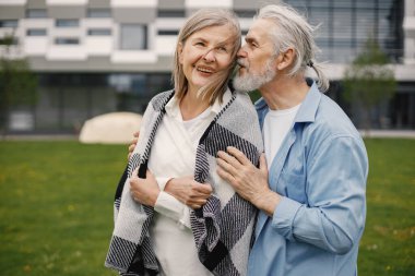 Yazın çimlerin üzerinde duran Kafkasyalı yaşlı çift. Erkek ve kadın sarılıyor. Beyaz gömlekli ve ekose battaniyeli bir kadın..