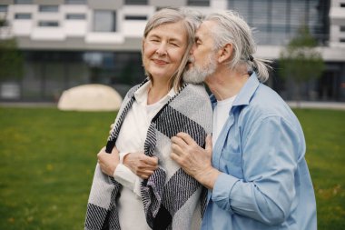 Yazın çimlerin üzerinde duran Kafkasyalı yaşlı çift. Erkek ve kadın sarılıyor. Beyaz gömlekli ve ekose battaniyeli bir kadın..