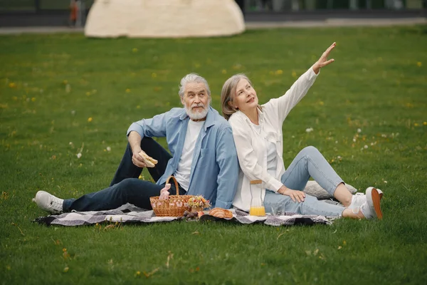Beautiful caucasian elderly couple sitting on a grass in the park in summer. Man and woman sitting on blanket at the park eating sandwiches and sharing few precious memories. Woman wearing white shirt
