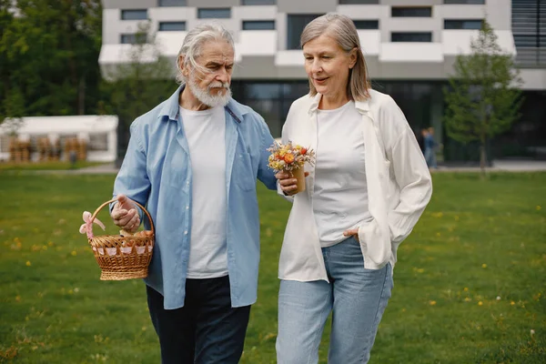 Caucasian elderly couple standing on a grass in summer. Man holding straw basket and woman flowers. Woman wearing white shirt and man blue one.