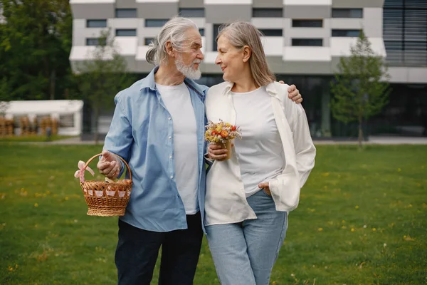 Caucasian elderly couple standing on a grass in summer. Man holding straw basket and woman flowers. Woman wearing white shirt and man blue one.