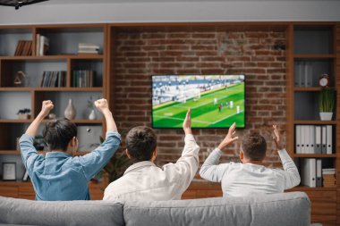 Bir futbol maçı izlemek için evde toplanan erkek arkadaşların arka görüntüsü. Erkekler büyük ekran televizyonun önünde kanepede oturuyor. Erkekler en sevdikleri takımı destekliyor ve maç hakkında yorum yapıyorlar.