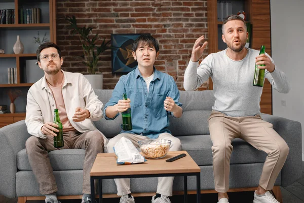 Front view of male friends gathered at home to watch a football match. Men sitting on the couch in front of the big screen TV and eating snacks. Men actively support their favorite team and feeling