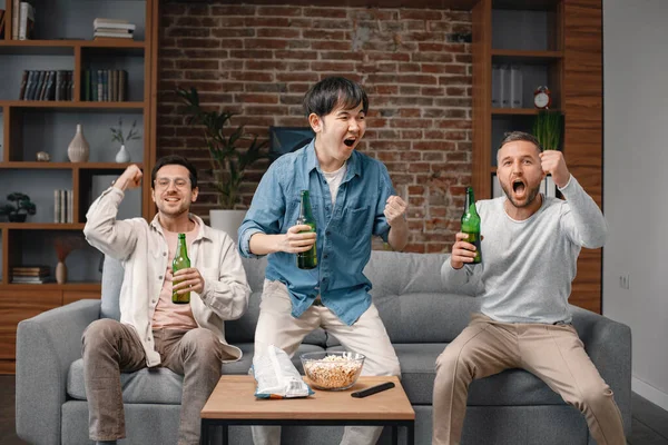 Front view of male friends gathered at home to watch a football match. Men sitting on the couch in front of the big screen TV and eating snacks. Men actively support their favorite team and feeling